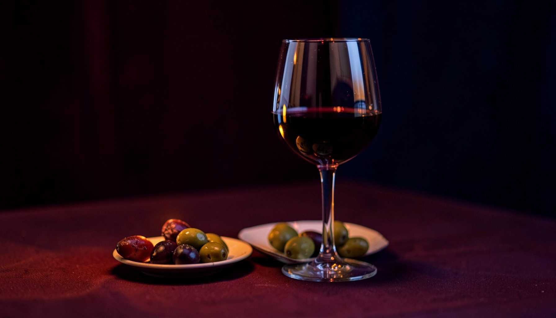 A glass of red wine beside a small plate of olives, candlelit table, deep burgundy and indigo mood