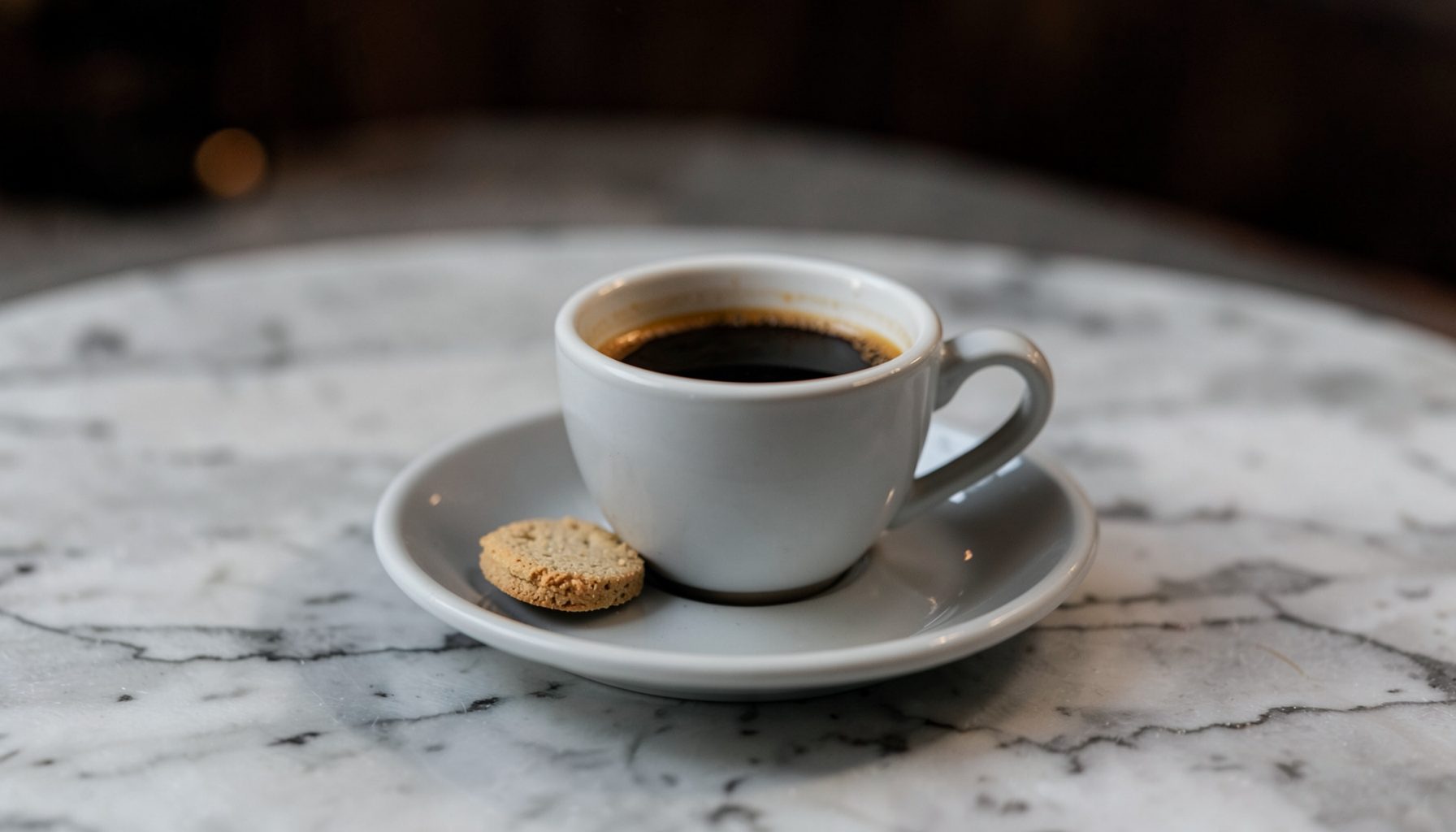 An espresso cup on a saucer with a small biscuit, marble tabletop, dark espresso brown and soft grey tones
